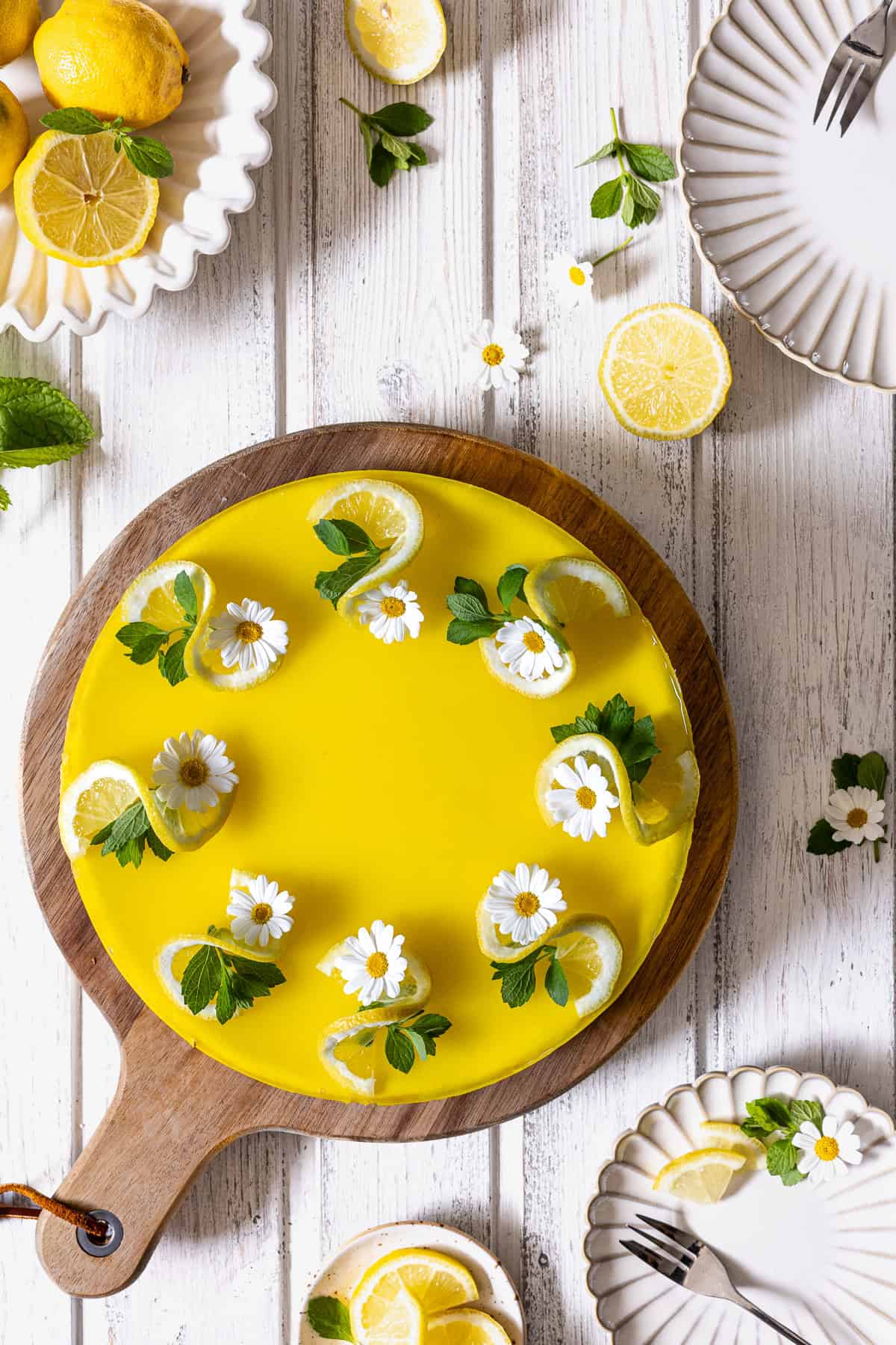 Ein cremiger Zitronenkäsekuchen mit knallgelbem Zitronenspiegel, dekoriert mit gewellten Zitronenscheiben, frischer Minze und weißen Margeriten-Blüten. Der Cheesecake liegt als ganzer Sommerkuchen auf einem runden Holzbrett mit Griff. Aufgenommen als Flatlay von oben auf einem hellen, weißen Holzhintergrund im Scandi-Style. Drumherum arrangiert sind weißes, geriffeltes Geschirr, Kuchengabeln, frische Zitronen in einer Schale und verstreute Minzblätter. Ein erfrischendes Rezept für den Sommer.