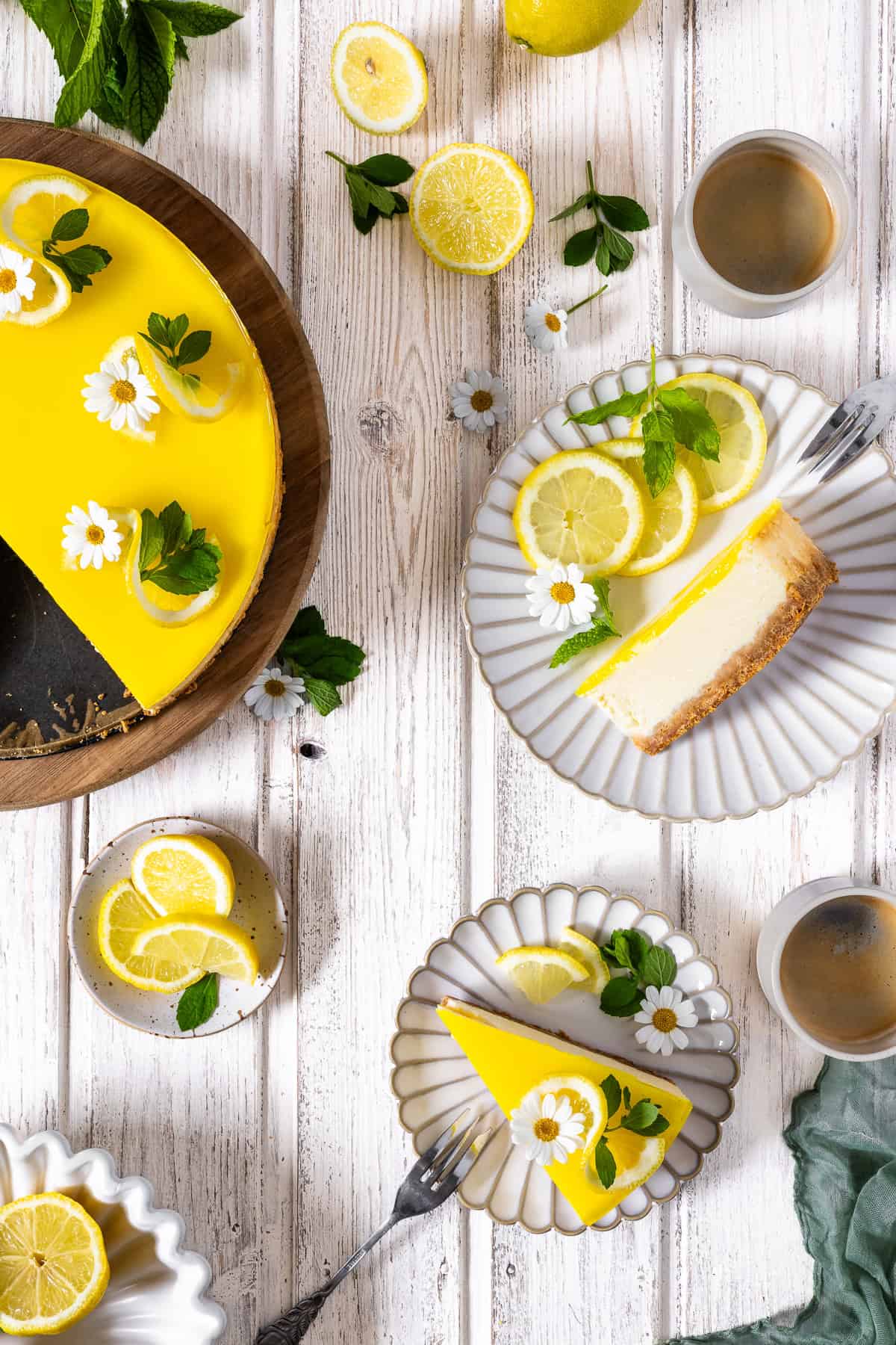 Ein angeschnittener Zitronenkäsekuchen mit spritzig-gelbem Zitronenspiegel auf einem runden Holzbrett. Auf zwei kleinen, geriffelten Keramiktellern daneben sind Tortenstücke angerichtet: Ein Cheesecake-Stück liegt flach auf der Seite und zeigt die cremigen Schichten aus Keksboden und Quarkmasse, das andere Stück steht aufrecht und ist mit Zitronenwellen, Minze und Gänseblümchen dekoriert. Das Flatlay auf weißem Holzhintergrund im hellen Scandi-Style wird durch zwei Tassen Kaffee, frische Zitronenscheiben und ein olivgrünes Leinentuch harmonisch ergänzt. Ein erfrischendes Sommerkuchen-Rezept.