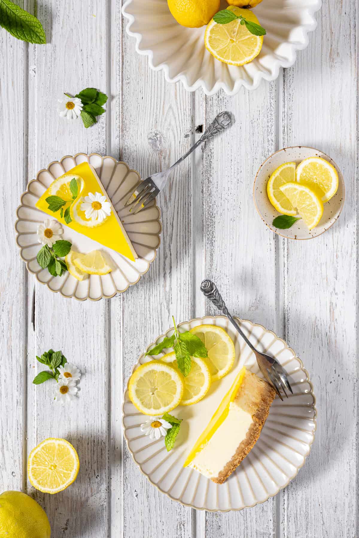 Zwei servierte Stücke Zitronenkäsekuchen auf weißen, geriffeltem Keramiktellern in der Draufsicht. Ein Cheesecake-Stück liegt flach auf der Seite und zeigt den Keksboden und die cremige Füllung, das andere Stück steht aufrecht mit glänzend gelbem Zitronenspiegel. Beide Stücke sind sommerlich mit Zitronenscheiben, frischer Minze und weißen Gänseblümchen dekoriert. Das Flatlay auf hellem, weißem Holzhintergrund wird durch Kuchengabeln, kleine Schälchen mit Zitronen und verstreute Blüten perfekt ergänzt. Ein erfrischendes Sommerkuchen-Rezept für die Kaffeetafel.