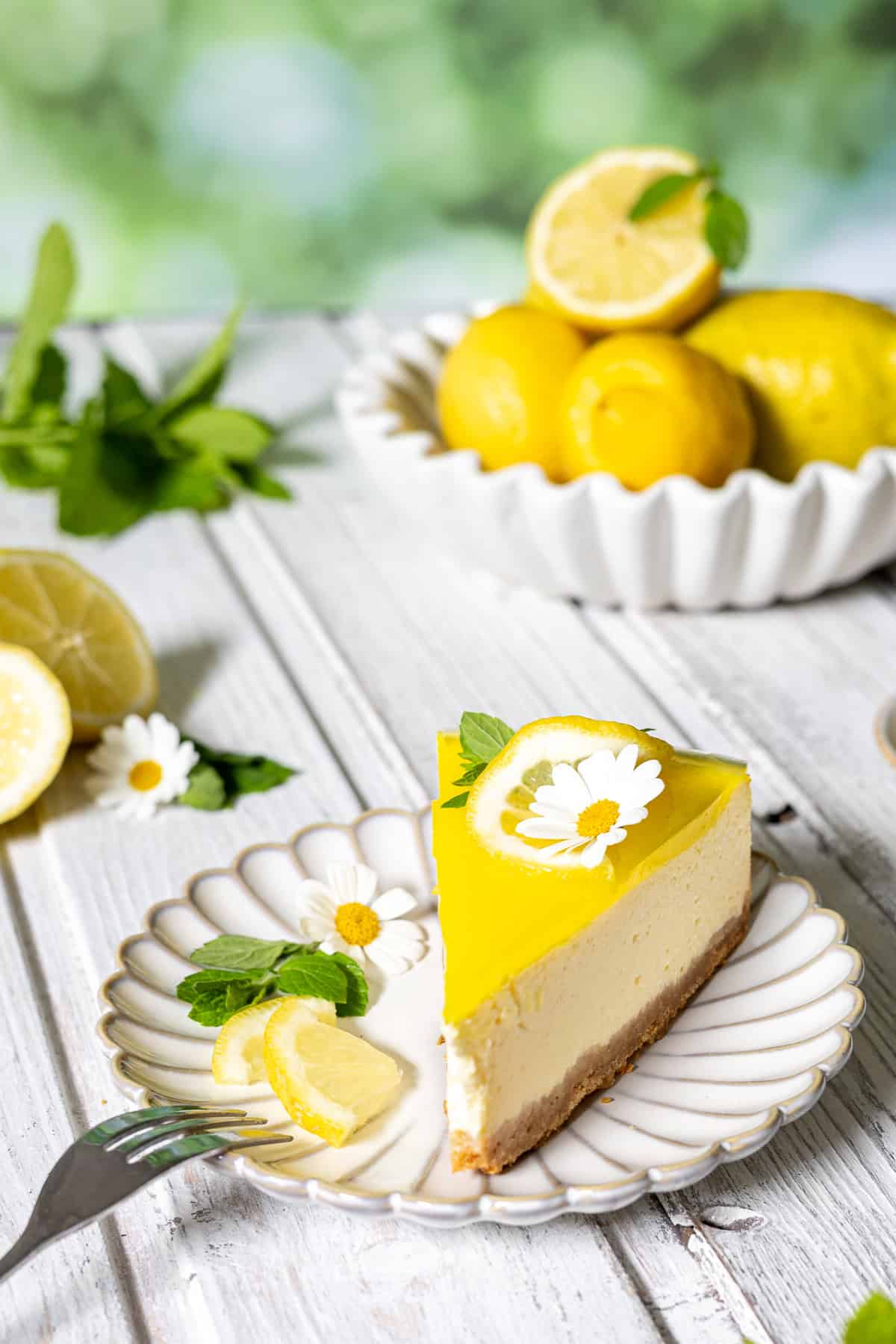 Nahaufnahme von einem cremigen Stück Zitronenkäsekuchen, aufrecht serviert auf einem weißen, geriffelten Keramikteller. Das Cheesecake-Stück zeigt perfekt die Schichten aus Keksboden, hoher Quarkmasse und gelbem Zitronenspiegel. Es ist dekoriert mit einer Zitronenscheibe, frischer Minze und einer weißen Margeriten-Blüte. Im unscharfen Hintergrund (Bokeh) auf dem hellen Holzboden stehen eine weiße Schale voller frischer Zitronen und Minzzweige vor einem sommerlich grünen Hintergrund. Eine Kuchengabel liegt im Vordergrund. Ideal als erfrischender Sommerkuchen.