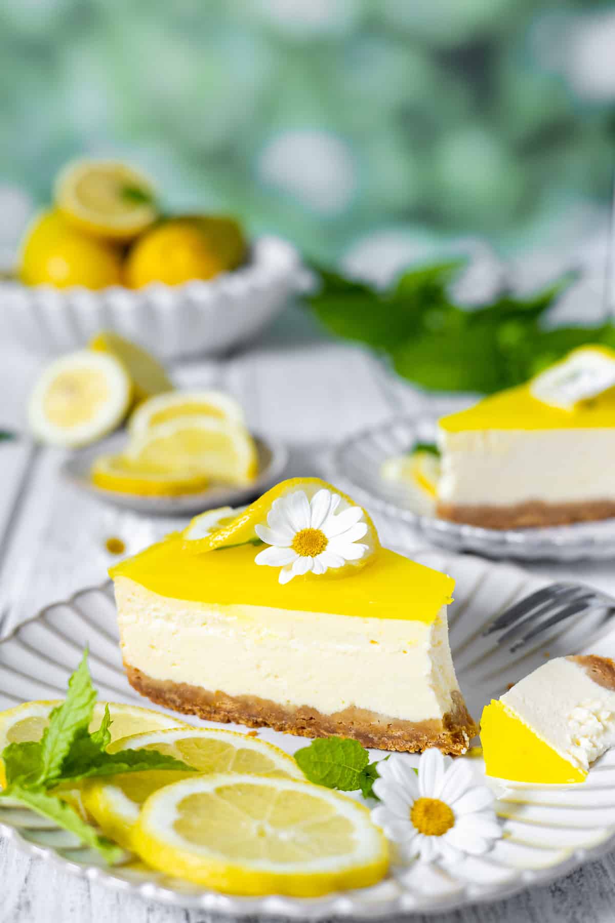 Detailaufnahme von einem cremigen Stück Zitronenkäsekuchen auf einem weißen, geriffelten Keramikteller aus der Augenhöhe-Perspektive. Das Cheesecake-Stück zeigt die samtige Quarkcreme auf einem knusprigen Keksboden und wird von einem spritzigen, gelben Zitronenspiegel gekrönt, der mit einer Zitronenwelle und einer weißen Margeriten-Blüte verziert ist. Ein kleines, abgeschnittenes Eckchen liegt dekorativ daneben. Im unscharfen Hintergrund sind ein weiteres Kuchenstück, frische Zitronenscheiben und eine weiße Schale vor einem weichen, grünen Sommer-Bokeh zu sehen. Ein erfrischendes Sommerkuchen-Rezept.