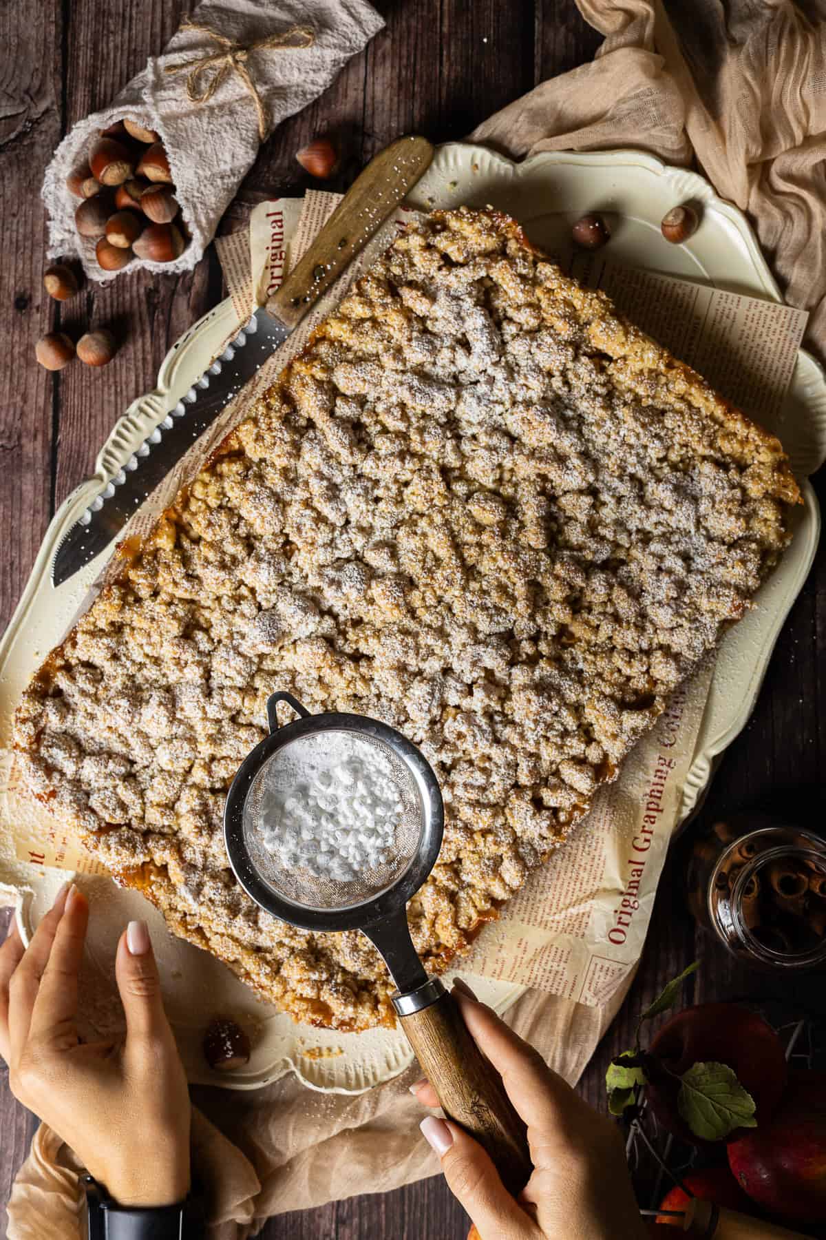 Ein üppiger, goldbrauner Apfelkuchen aus dem Thermomix®, im Ganzen als Blechkuchen auf einer großen Servierplatte mit bedrucktem Vintage-Papier angerichtet. Zwei Hände sind im Bild zu sehen: Eine bestäubt die knusprigen Streusel gerade in Aktion mit einem Puderzuckersieb. Ein langes Messer mit Holzgriff und herbstliche Deko runden das authentische Back-Szenario von oben ab.