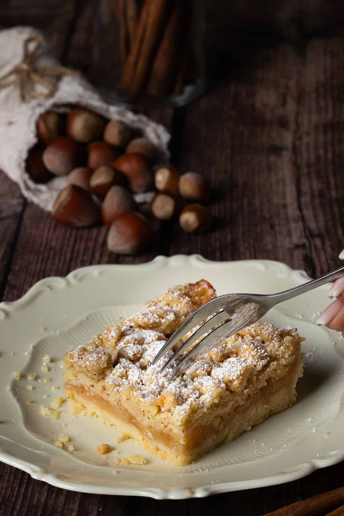 Ein Stück saftiger Apfelkuchen mit dicken Nuss-Streuseln und Puderzucker, angerichtet auf einem cremefarbenen Vintage-Teller auf einem dunklen Holztisch. Eine Hand sticht mit einer silbernen Kuchengabel in die weiche, fruchtige Apfelschicht. Im gemütlichen, unscharfen Hintergrund liegt ein rustikales Leinensäckchen, aus dem ganze Haselnüsse herauskullern.