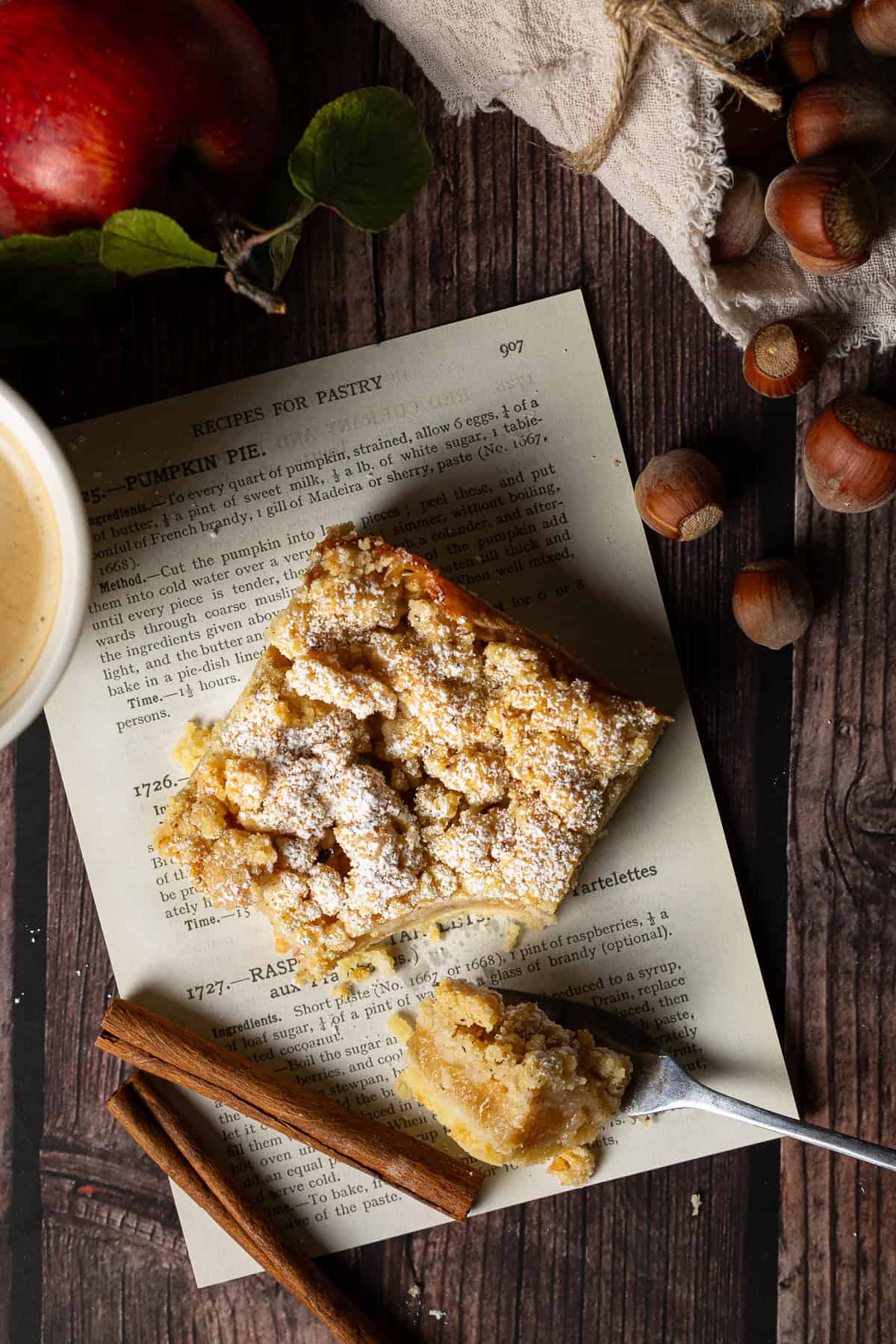 Ein gemütlicher Herbstkuchen mit Äpfeln und Streuseln, rustikal serviert auf bedrucktem Vintage-Papier. Links im Bild ist der Rand einer weißen Kaffeetasse angeschnitten. Neben dem saftigen Kuchenstück liegt eine antike Gabel mit einem kleinen Bissen, flankiert von Zimtstangen, einem Haselnuss-Säckchen und leuchtend roten Äpfeln für die perfekte Herbststimmung.